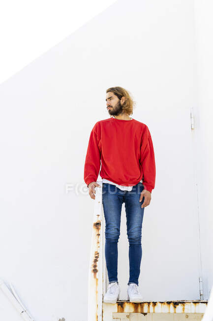 Retrato de un joven barbudo con sudadera roja de pie en escaleras oxidadas - foto de stock