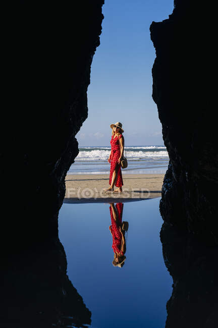 Blonde Frau mit rotem Kleid und Hut am Strand entlang, Naturbogen an der Playa de Las Catedrales, Spanien — Stockfoto