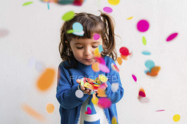 Feliz niña soplando el confeti en una fiesta frente a una pared blanca - foto de stock