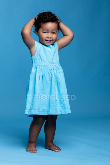 Retrato de niña sonriente con las manos en la cabeza usando vestido azul claro de pie sobre fondo azul - foto de stock