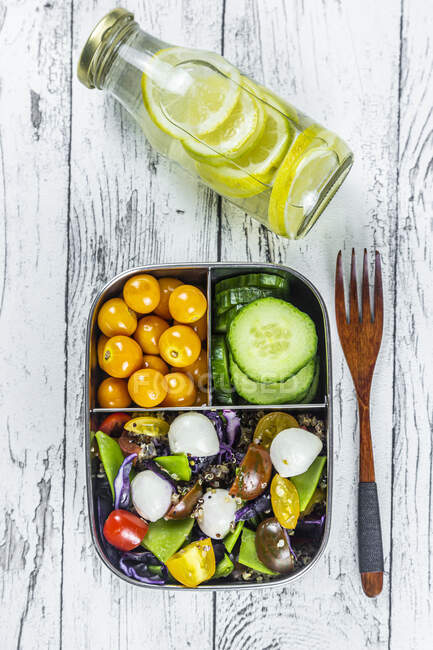 Bottle of lemonade and lunchbox with cucumber slices, winter cherries and quinoa salad (quinoa, cherry tomato, red cabbage, sugar snap peas and mozzarella balls) — Stock Photo