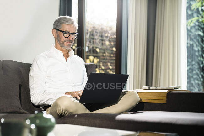 Senior man with grey hair in modern design living room working on laptop in home office — Stock Photo