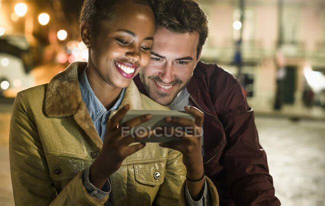 Портрет счастливой молодой пары, смотрящей вместе на смартфон ночью, Лиссабон, Португалия — стоковое фото