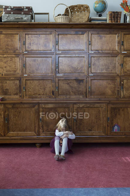 Niña infeliz sentada en el suelo frente al gabinete grande - foto de stock