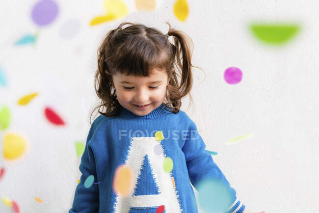 Niña feliz con el confeti durante una fiesta frente a una pared blanca - foto de stock