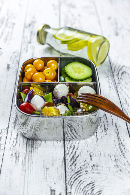 Bottle of lemonade and lunchbox with cucumber slices, winter cherries and quinoa salad (quinoa, cherry tomato, red cabbage, sugar snap peas and mozzarella balls) — Stock Photo