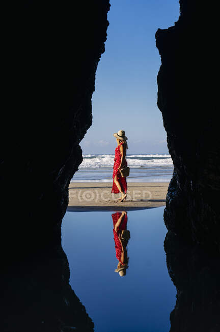 Жінка-блондинка у червоній сукні і капелюсі і йде уздовж пляжу, Natural Arch at Playa de Las Catedrales, Spain — стокове фото