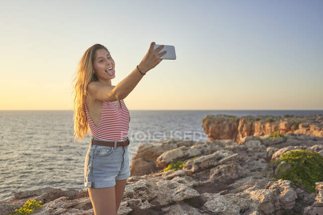 Jovem mulher de pé no penhasco no mar, usando smartphone, tirando selfie — Fotografia de Stock