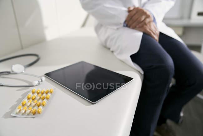Doctor sitting on table in his medical practice with tablet, stethoscope and capsules — Stock Photo