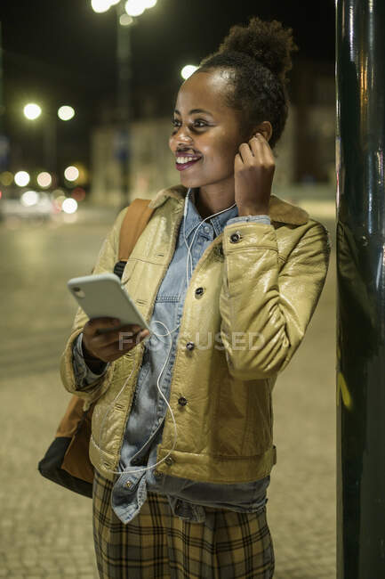 Ritratto di giovane donna sorridente che usa cuffie e smartphone in città di notte, Lisbona, Portogallo — Foto stock