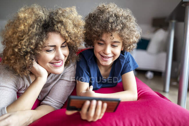 Madre e figlio guardando un video su smartphone, sdraiati su un grande cuscino — Foto stock