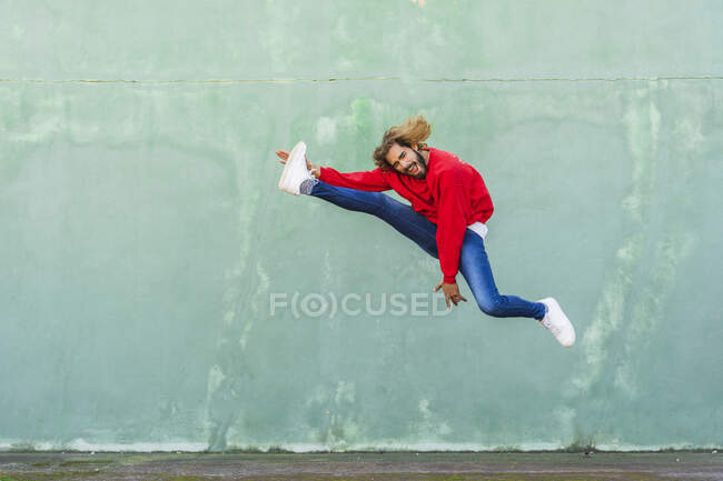 Retrato de un joven con sudadera roja saltando en el aire frente a la pared verde - foto de stock