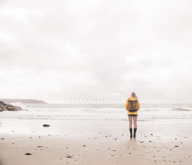 Позаду жінка в жовтому дощовому піджаку стоїть на пляжі. — стокове фото