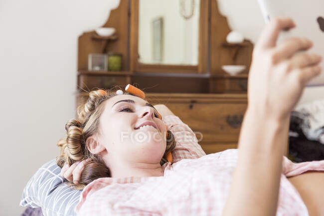 Young woman with curlers in hair lying on bed taking selfie with digital tablet — Stock Photo