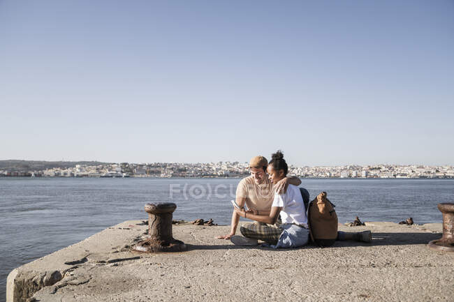 Young couple sitting on pier at the waterfront using cell phone, Lisbon, Portugal — Stock Photo