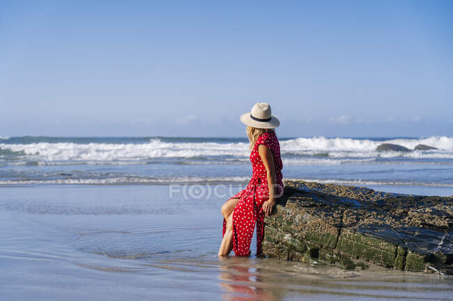 Blond woman wearing red dress and hat sittig on rock at the beach, Playa de Las Catedrales, Spain — Stock Photo