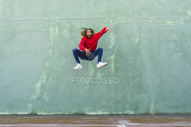 Портрет молодого чоловіка в червоній сорочці, що стрибає в повітрі перед зеленою стіною — стокове фото
