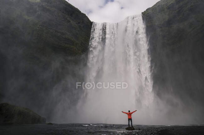 Hombre maduro parado frente a las cascadas de Skogafoss con los brazos levantados, Islandia - foto de stock