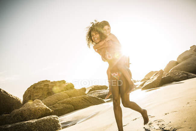 Happy mother carrying her son piggyback on the beach — Stock Photo