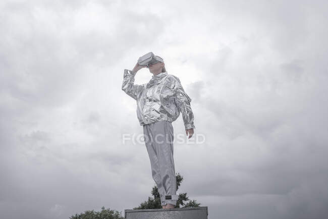 Girl in silver suit looking through VR goggles, standing on box, against grey sky — Stock Photo