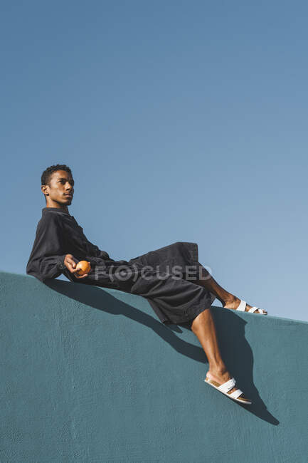 Молодой человек в черном кафтане лежит на голубой стене с апельсинами — стоковое фото