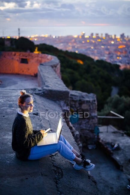 Young woman sitting on railing above the city using cell phone, Barcelona, Spain — Stock Photo