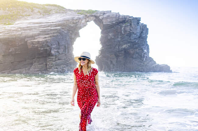 Жінка-блондинка у червоній сукні і капелюсі і йде уздовж пляжу, Natural Arch at Playa de Las Catedrales, Spain — стокове фото