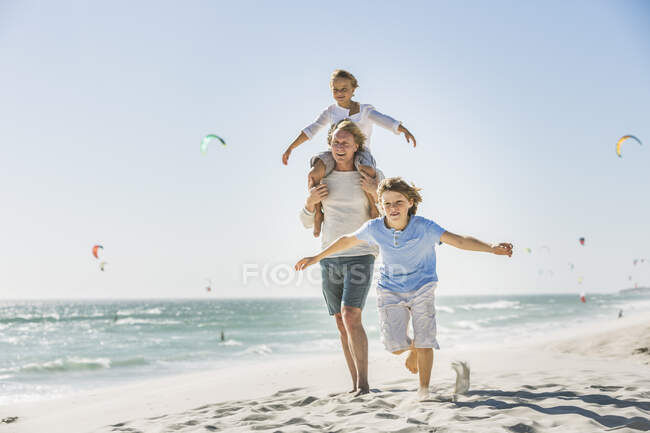 Padre divirtiéndose con sus hijos en la playa, llevando uno a cuestas - foto de stock