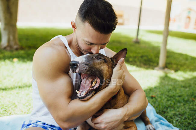 Muscular man sitting on blanket on a meadow hugging his dog — Stock Photo