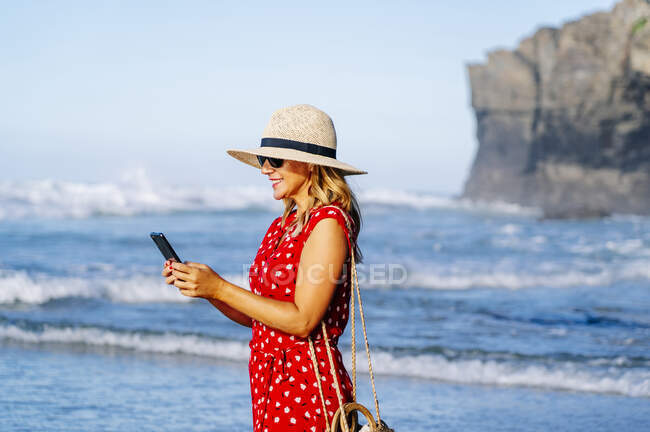 Femme blonde portant une robe rouge et un chapeau et utilisant un smartphone à la plage — Photo de stock