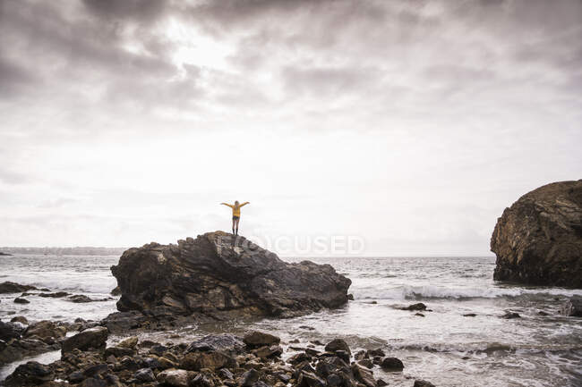 Позаду жінка з піднятими руками стоїть на скелястому пляжі. — стокове фото