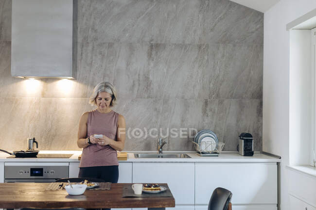 Зріла жінка використовує мобільний телефон на кухні вдома — стокове фото