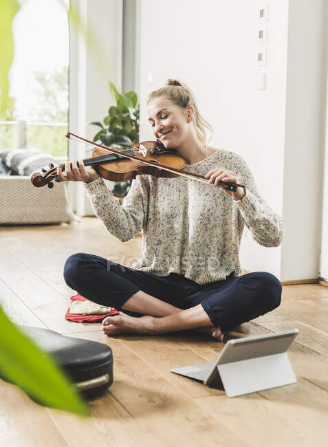 Жінка з планшетом сидить вдома на підлозі, граючи на скрипці. — стокове фото