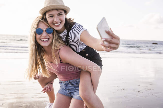 Deux petites amies s'amusent sur la plage, se portent mutuellement, prennent des selfies de smartphone — Photo de stock