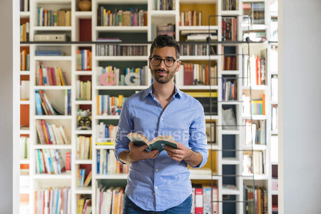 Portrait of smiling young man with book standing in front of bookshelves at home — Stock Photo