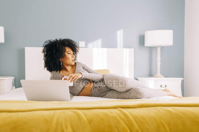 Молода жінка з чашкою кави і ноутбуком лежить на ліжку вдома — стокове фото