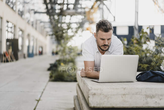 Young man with laptop and looking sideways, lying on a bench in the city — Stock Photo