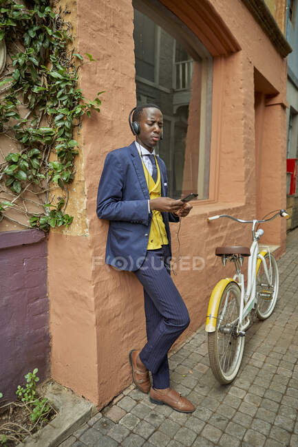 Стильный молодой бизнесмен с велосипедом в старомодном костюме слушает музыку на наушниках — стоковое фото