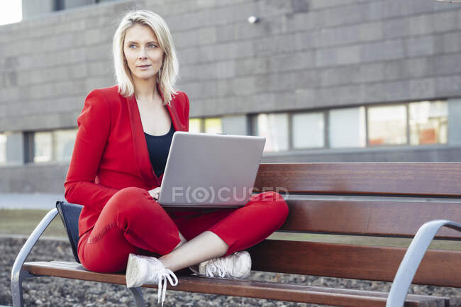 Mujer de negocios rubia con traje rojo y portátil en un banco - foto de stock
