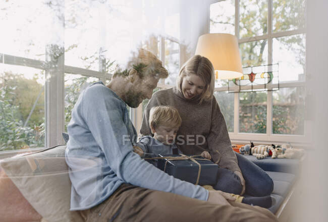 Маленький мальчик с родителями разворачивает подарок в солнечной комнате дома — стоковое фото
