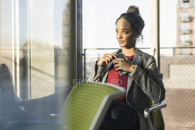 Молода бізнесменка з мобільним телефоном сидить біля вікна в офісі. — стокове фото