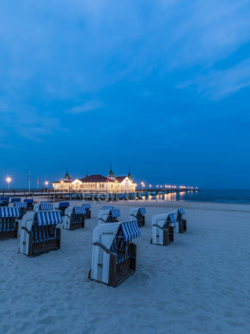 Germania, Meclemburgo-Pomerania occidentale, Heringsdorf, Sedie da spiaggia con cappuccio sulla spiaggia sabbiosa costiera al crepuscolo con molo illuminato sullo sfondo — Foto stock