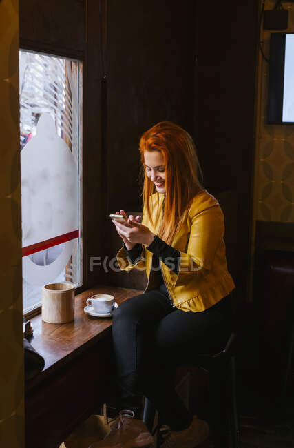 Happy redheaded young woman sitting in a coffee shop using cell phone — Stock Photo