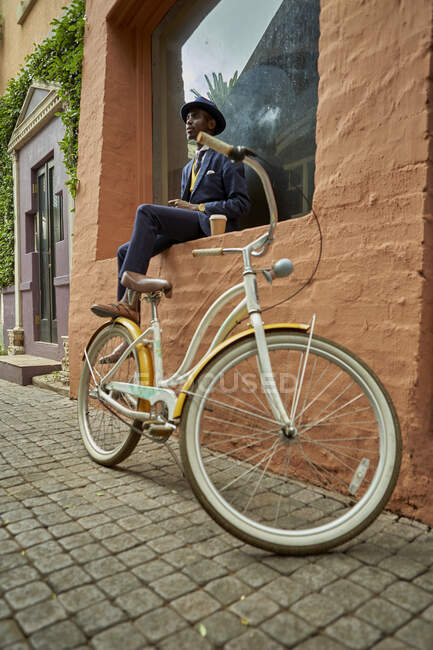 Elegante joven hombre de negocios con bicicleta con traje anticuado sentado en una repisa de una ventana - foto de stock