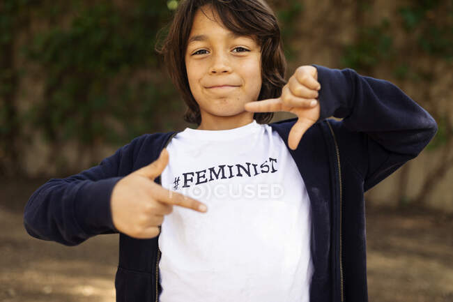 Niño pequeño parado en la calle con estampado en camiseta, diciendo Feminista, haciendo marco de dedo - foto de stock