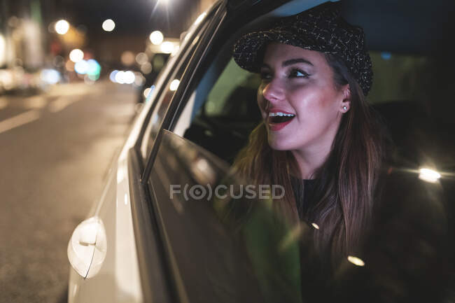 Mujer sentada en el asiento trasero de un coche en la ciudad por la noche, mirando por la ventana del coche - foto de stock