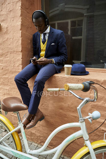 Elegante giovane uomo d'affari con bicicletta che indossa un abito vecchio stile seduto su un cornicione di una finestra ad ascoltare musica sulle cuffie — Foto stock