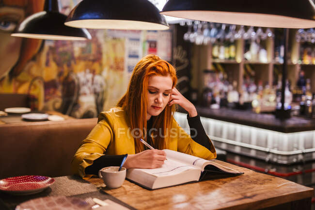 Retrato de una joven pelirroja a la mesa en un pub escribiendo en un libro - foto de stock