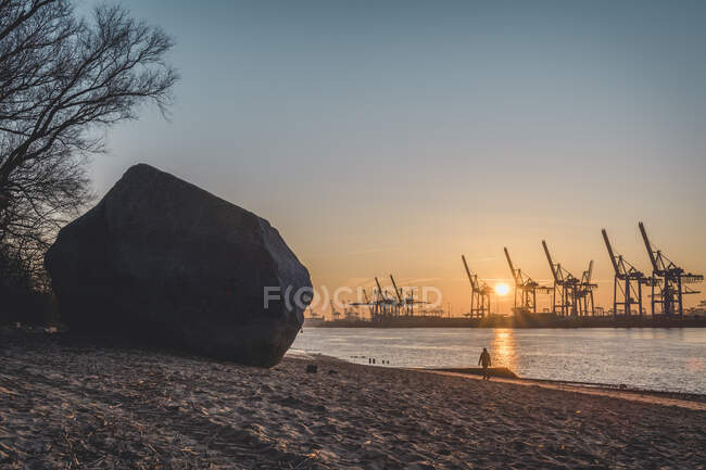 Alemanha, Hamburgo, Alter Schwede pedregulho na praia ribeirinha ao nascer do sol com silhuetas de guindastes portuários no fundo — Fotografia de Stock