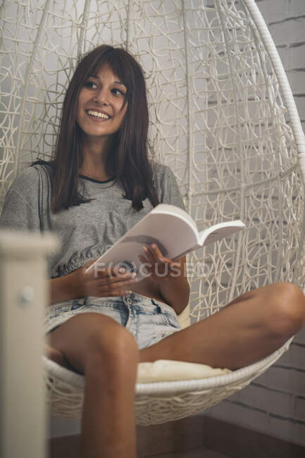Lächelnde junge Frau sitzt im Hängesessel und liest ein Buch — Stockfoto